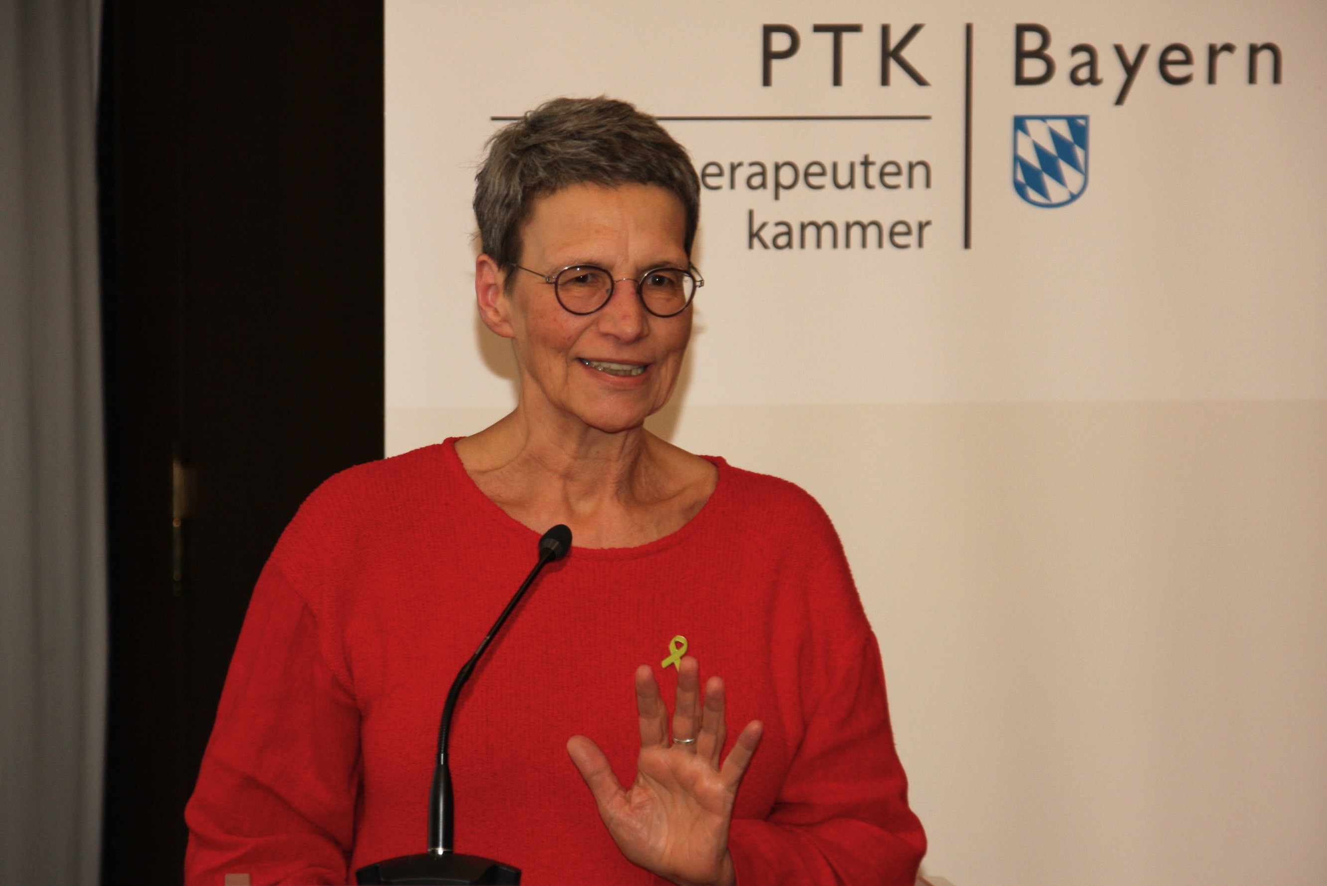 Eine Frau mit rotem Oberteil und Brille steht am Redepult. Im Hintergrund ist ein Banner der PTK Bayern zu sehen.